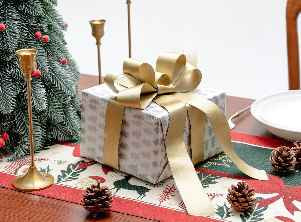 Christmas gift wrapped in silver paper with a gold ribbon bow on a festive table runner with pinecones and a tree.