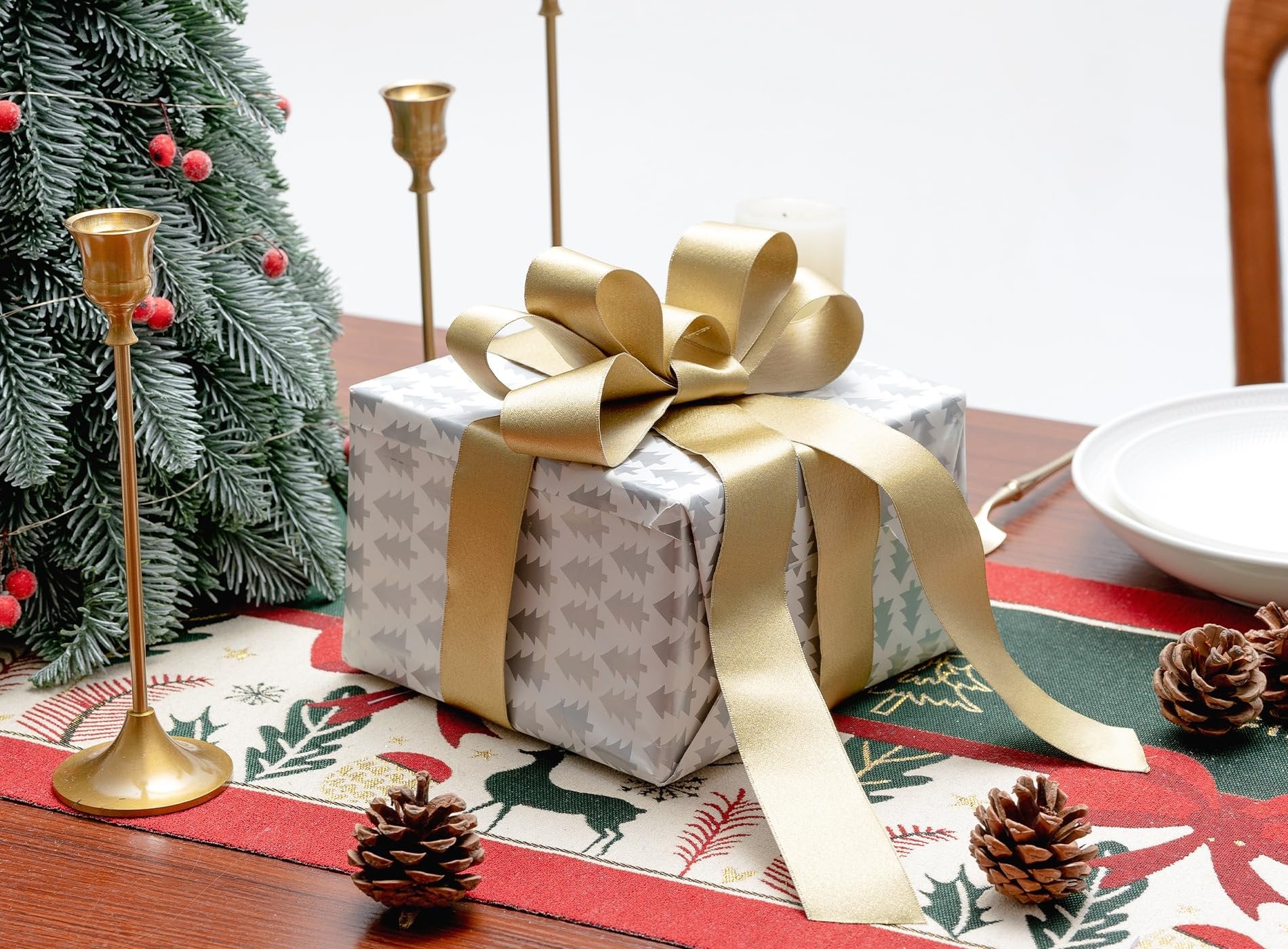 Christmas gift wrapped in silver paper with a gold ribbon bow on a festive table runner with pinecones and a tree.