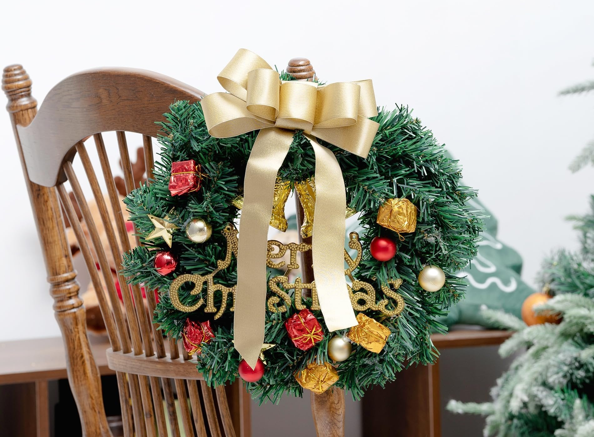 Christmas wreath with gold bow and 'Merry Christmas' text, decorated with ornaments and gift boxes, hanging on a wooden chair.