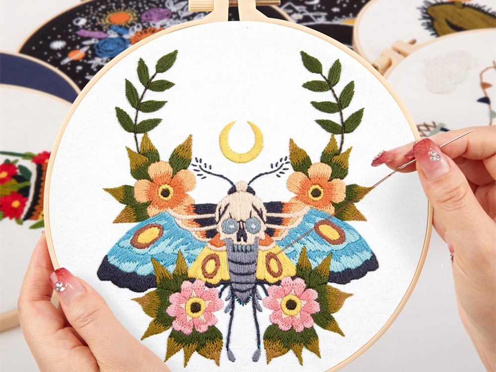 Embroidery hoop with a skull moth and floral pattern being stitched by a hand holding a needle.