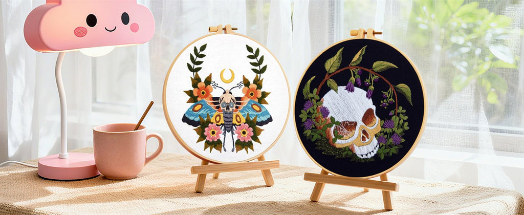 Two embroidery hoops with gothic skull and moth patterns displayed on wooden stands next to a pink lamp and mug.