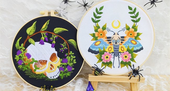 Two embroidery hoops with gothic designs: one with a skull and flowers on black fabric, the other with a moth and flowers on white fabric.