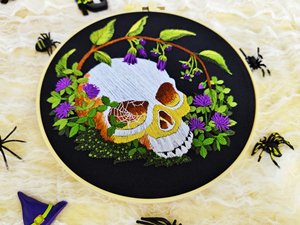 Embroidery hoop with a gothic skull and floral pattern, surrounded by Halloween decorations.