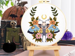 Embroidery hoop with gothic skull and moth pattern, displayed on a wooden easel with Halloween decorations.