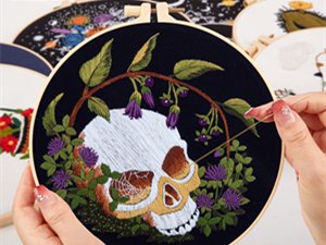Embroidery hoop with skull, flowers, and moth pattern being stitched by hands, multiple hoops in background