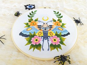 Embroidery hoop with skull moth and floral pattern, surrounded by decorative spiders and an evil eye patch.