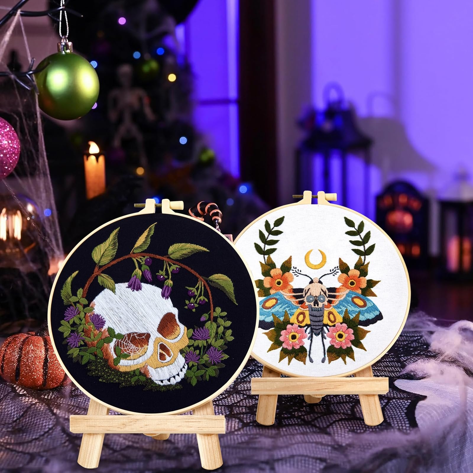 Two embroidery hoops with gothic skull and moth patterns displayed on wooden stands, with festive decorations in the background.