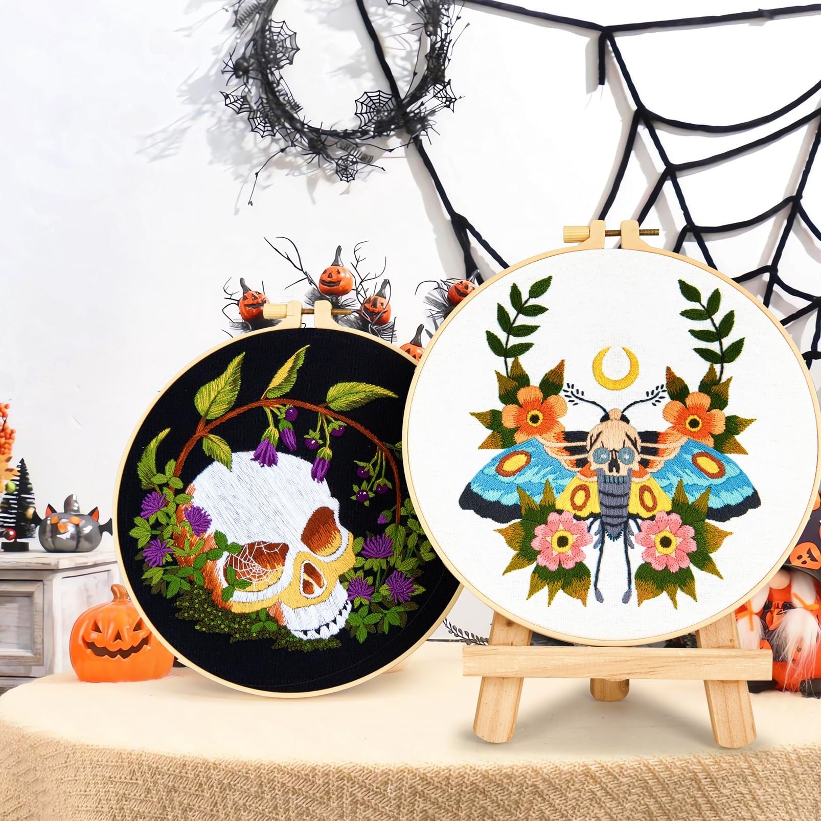 Two embroidery hoops, one with a skull and flowers on black fabric, the other with a moth and flowers on white fabric, displayed with Halloween decorations.