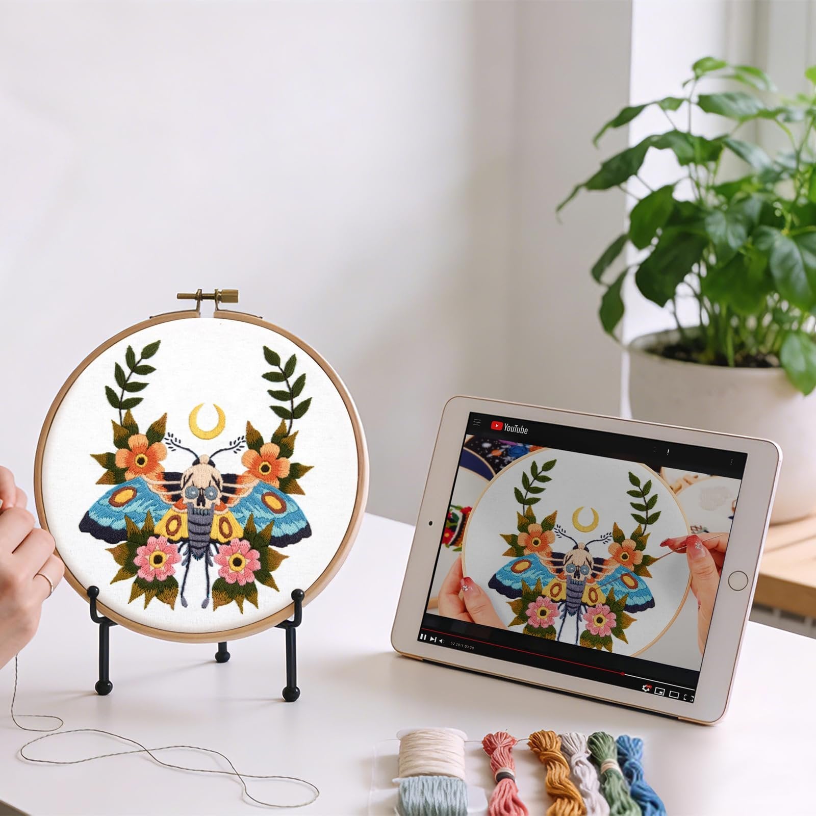 Embroidery hoop with moth skull pattern and tablet showing video tutorial on a white table with thread spools