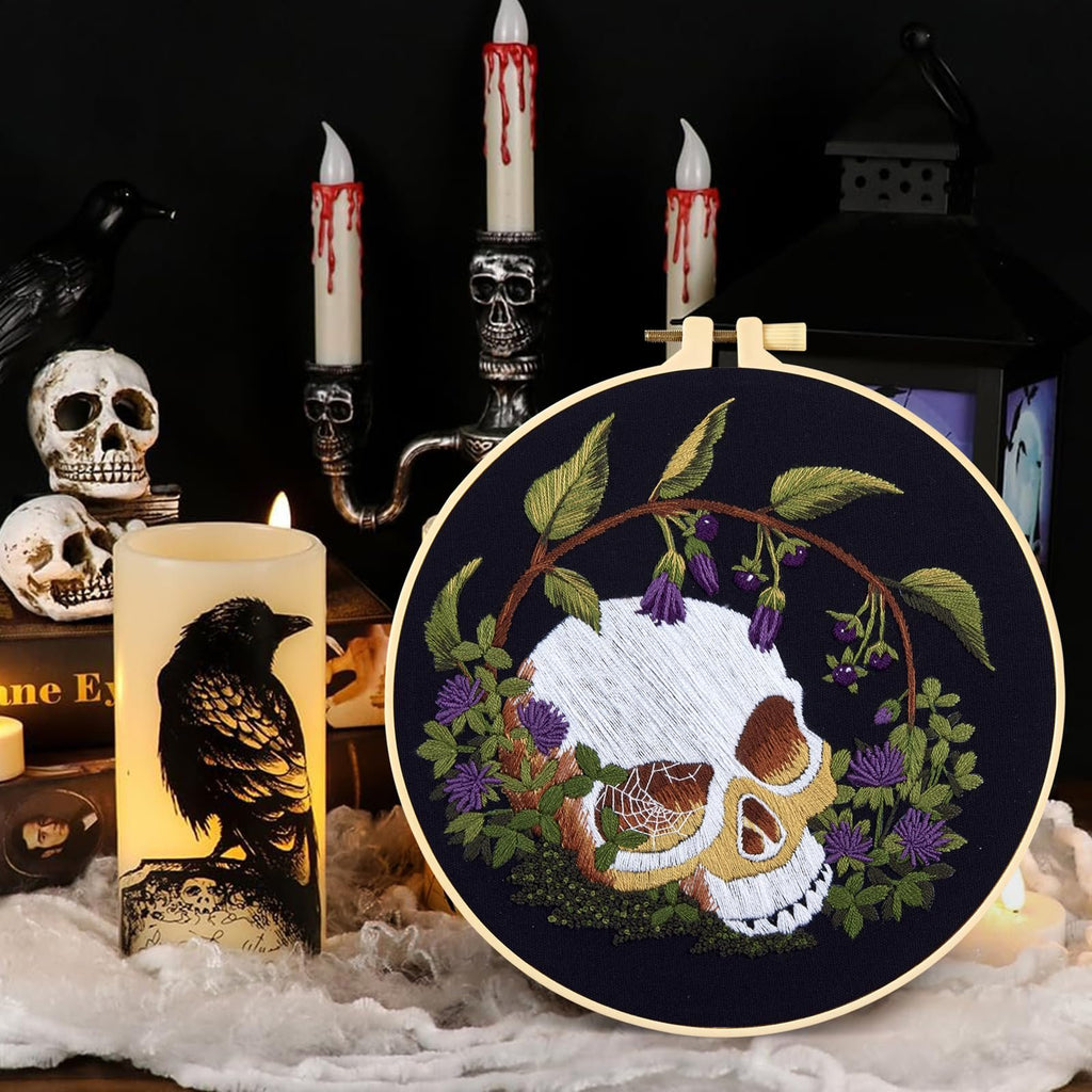 Embroidery hoop with a gothic skull and floral design, surrounded by Halloween-themed decor including skulls and candles.