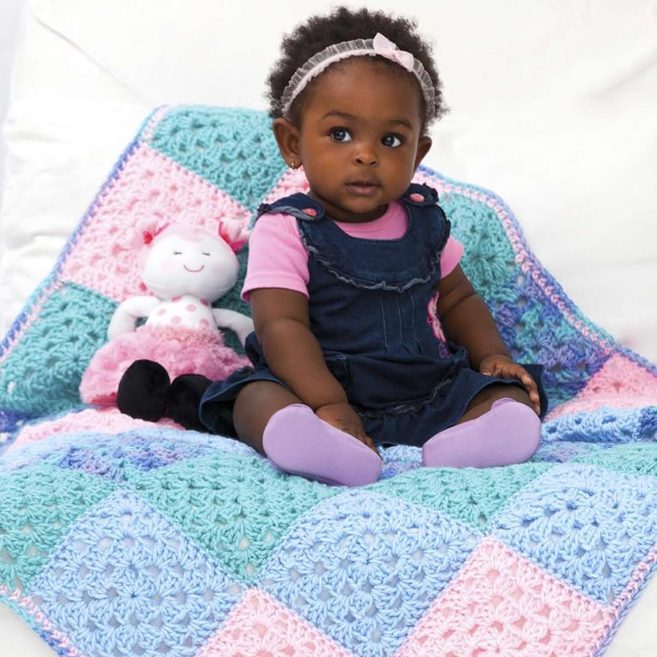Baby girl sitting on a colorful crocheted blanket with a stuffed doll.