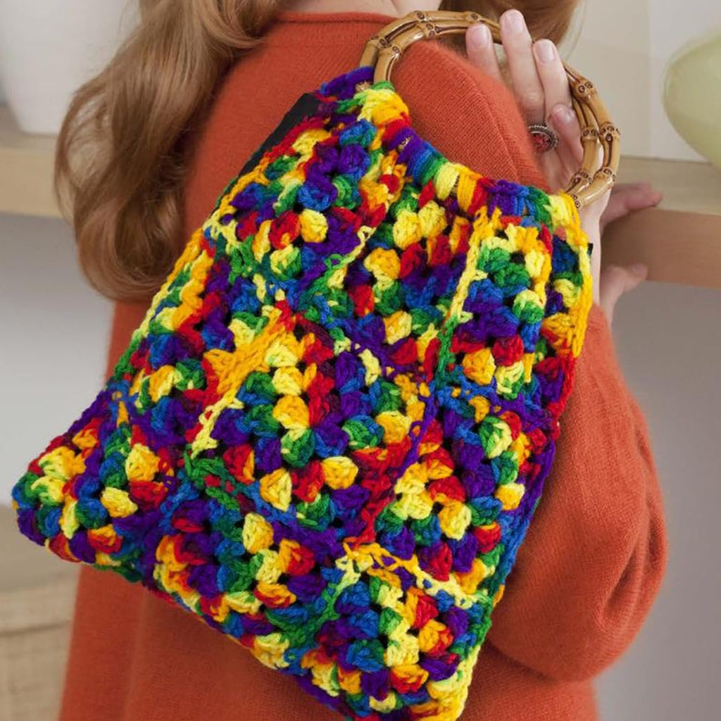 Woman wearing an orange sweater holding a colorful crocheted granny square handbag with bamboo handles.
