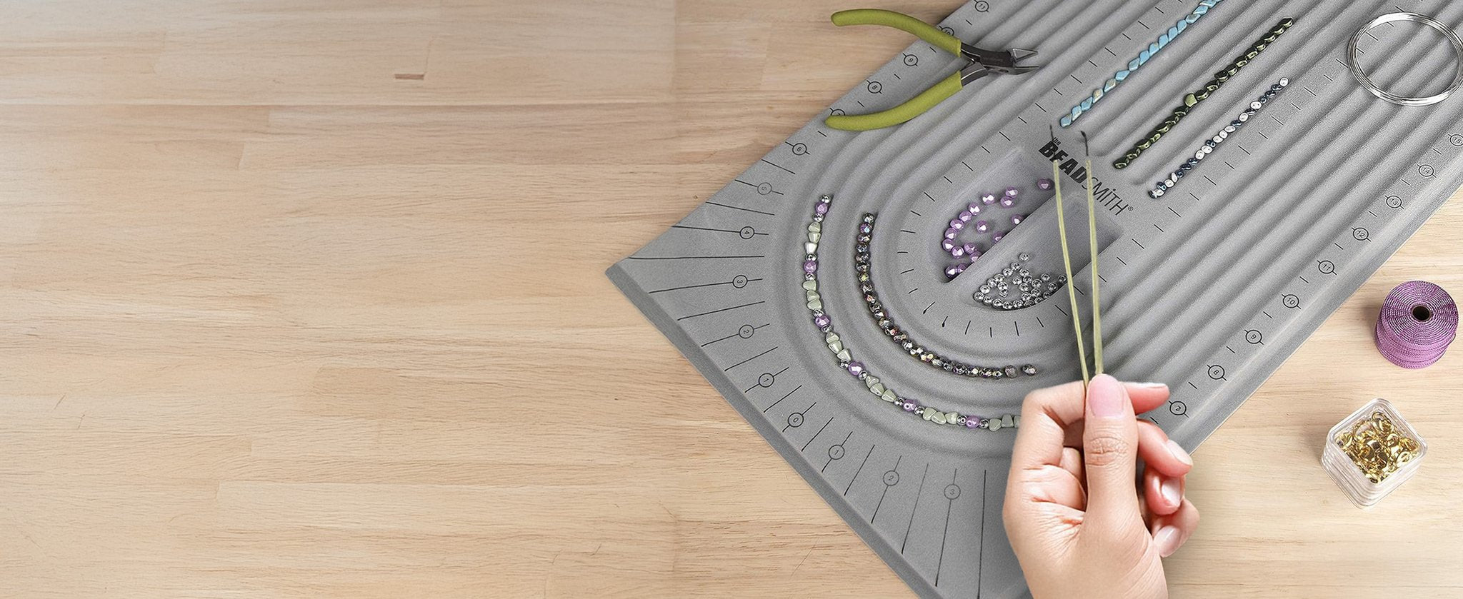 Grey flocked jewelry bead board with beads, tweezers, pliers, and thread on a wooden surface.