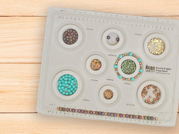 Grey flocked jewelry design board with compartments filled with beads and a bracelet, showing ruler markings.