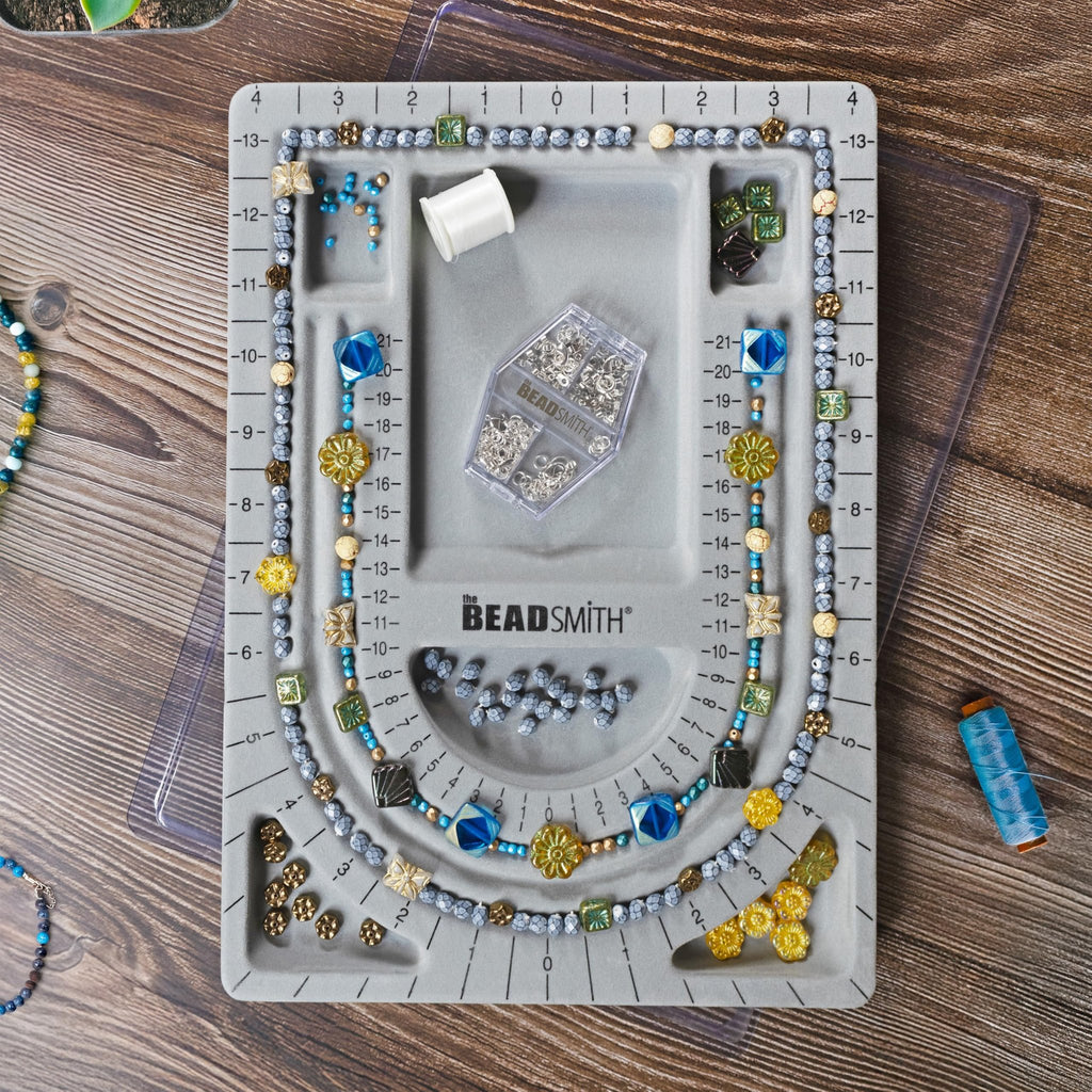Grey flocked jewelry bead board with beads and findings arranged on it, top-down view. Logo is visible on the board.