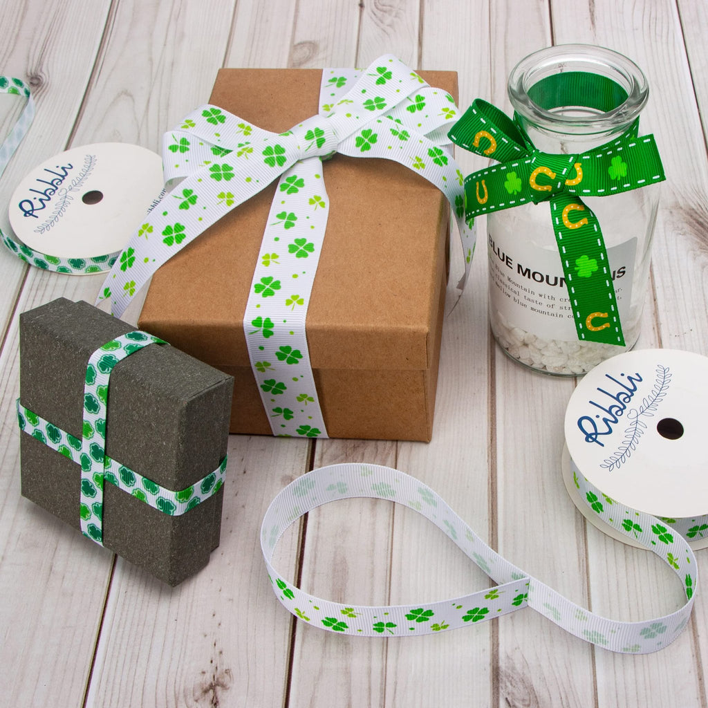 Ribbon with shamrocks and white dots on a spool, tied around a gift box and a dark gray box, and a glass jar with green ribbon.
