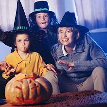 Three people in Halloween costumes with fake teeth and witch hats pose with a carved pumpkin.