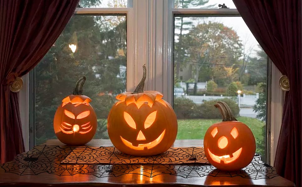 Three carved Halloween pumpkins glowing on a spider web table runner in front of a window with dark red curtains.