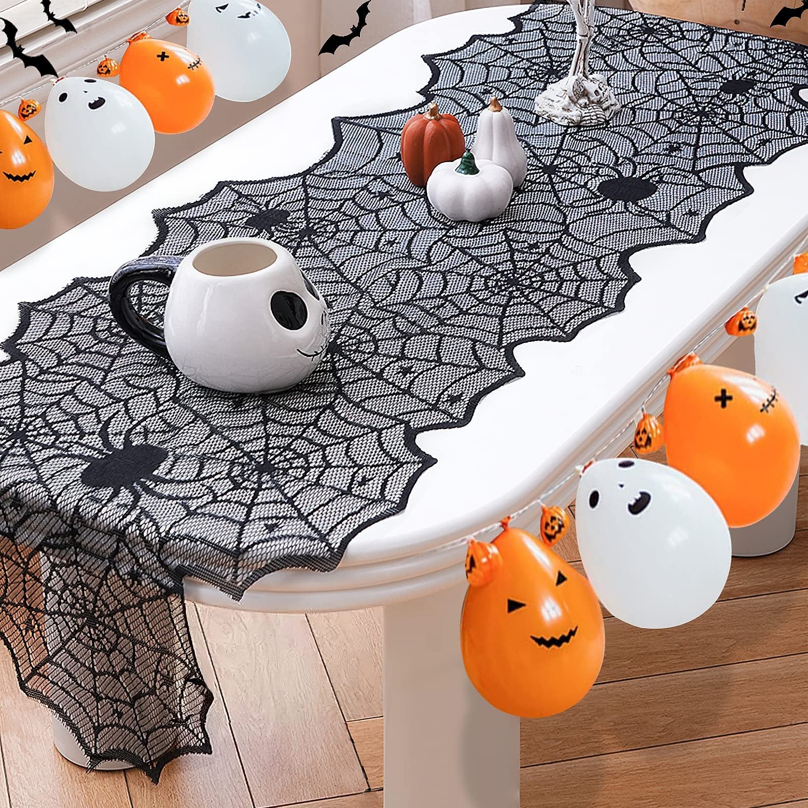 Black lace spider web table runner with spiders and pumpkins on a white table, decorated with Halloween balloons and bats.