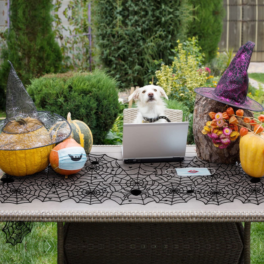 Halloween black lace spider web table runner on a table with pumpkins, witch hats, and a dog using a laptop.