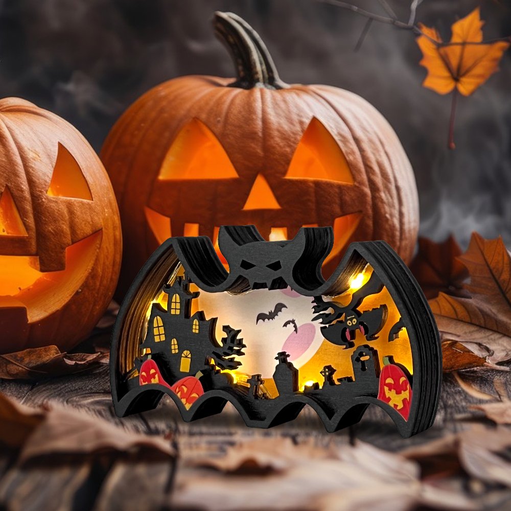 Halloween light-up bat shadow box with spooky scene, flanked by carved pumpkins on a wooden surface.