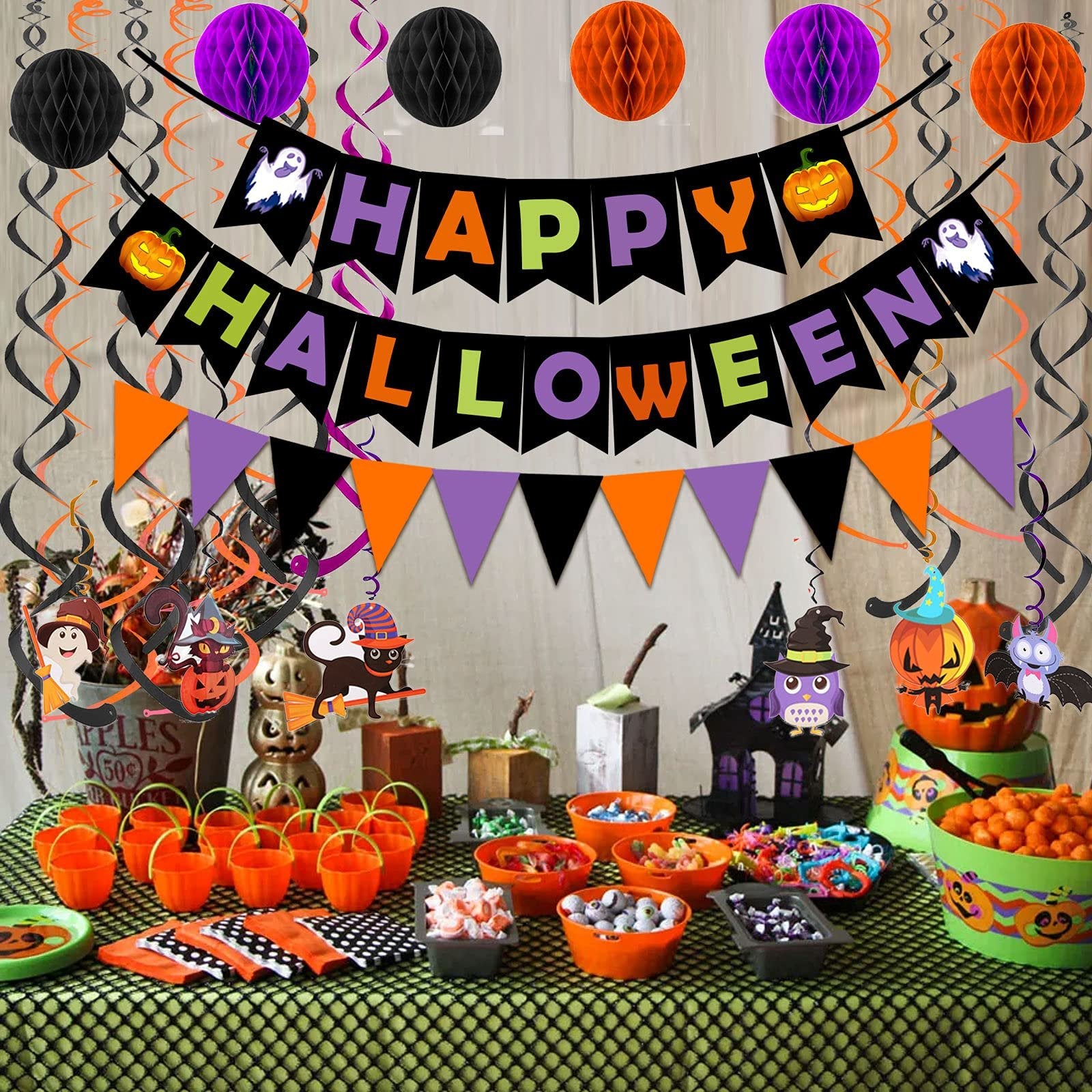 Halloween party table setup with decorations including a 'Happy Halloween' banner, honeycomb balls, swirl streamers, candy buckets, and themed candy bowls.