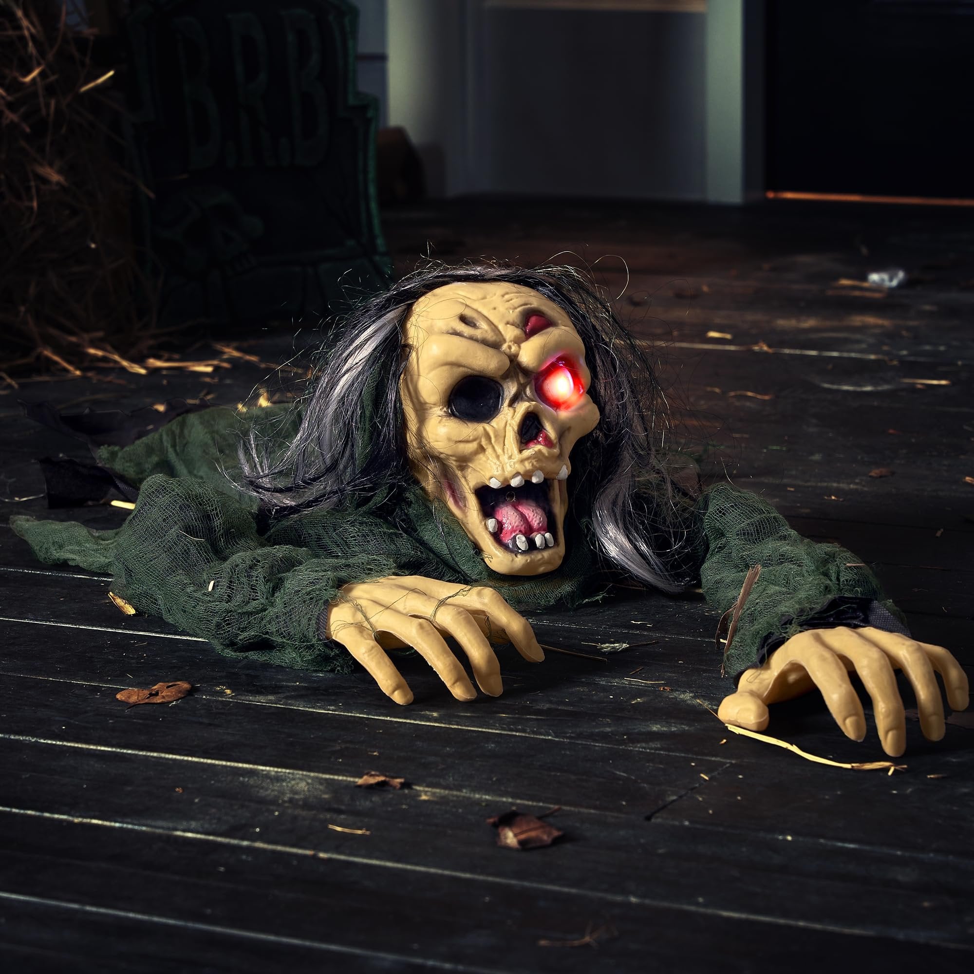 Scary zombie groundbreaker prop with glowing red eyes and tattered green clothing crawling on a wooden floor, with a tombstone in the background.