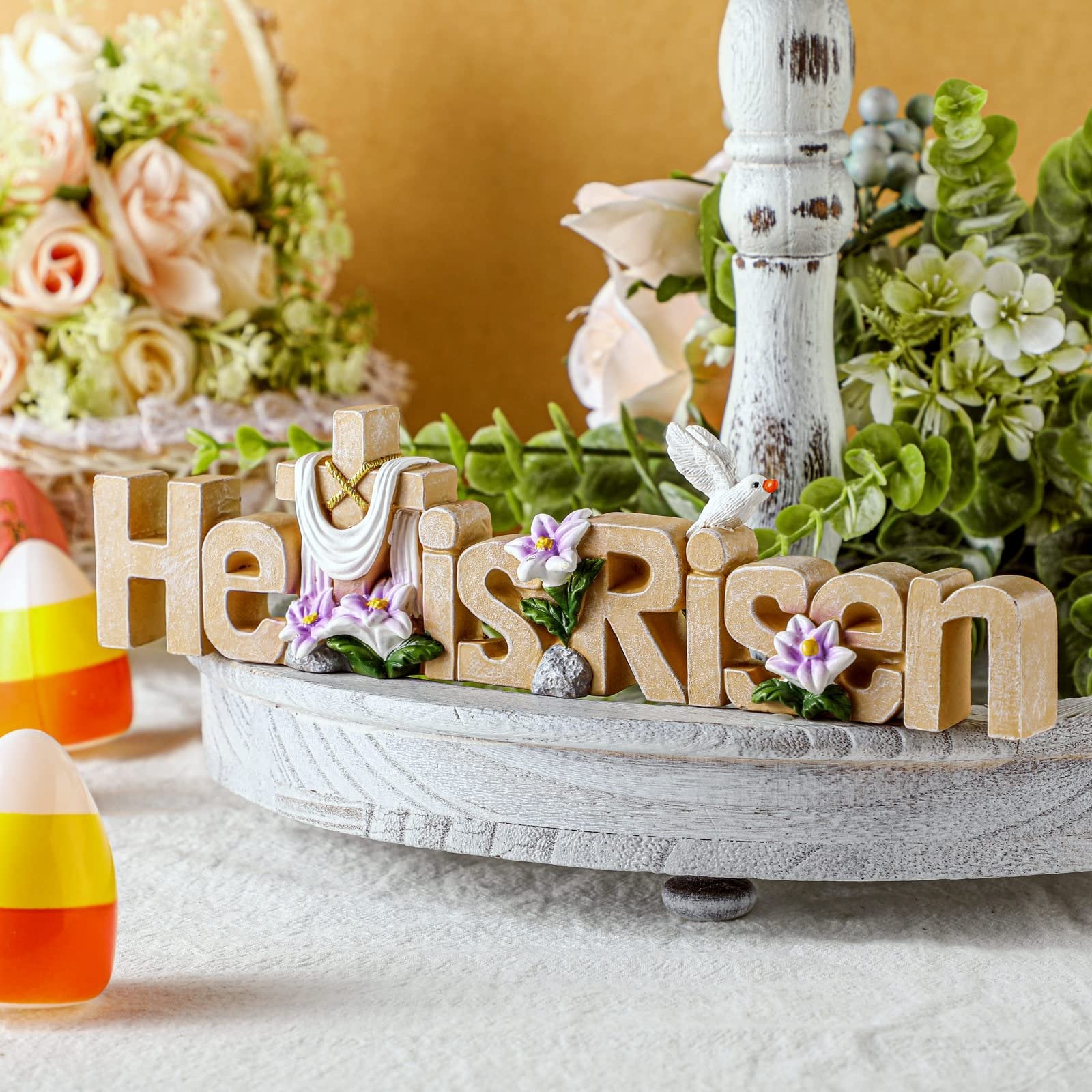 Easter tabletop sign with 'He is Risen' text, cross, dove, and flowers, displayed on a tiered tray with floral arrangements and candy corn.