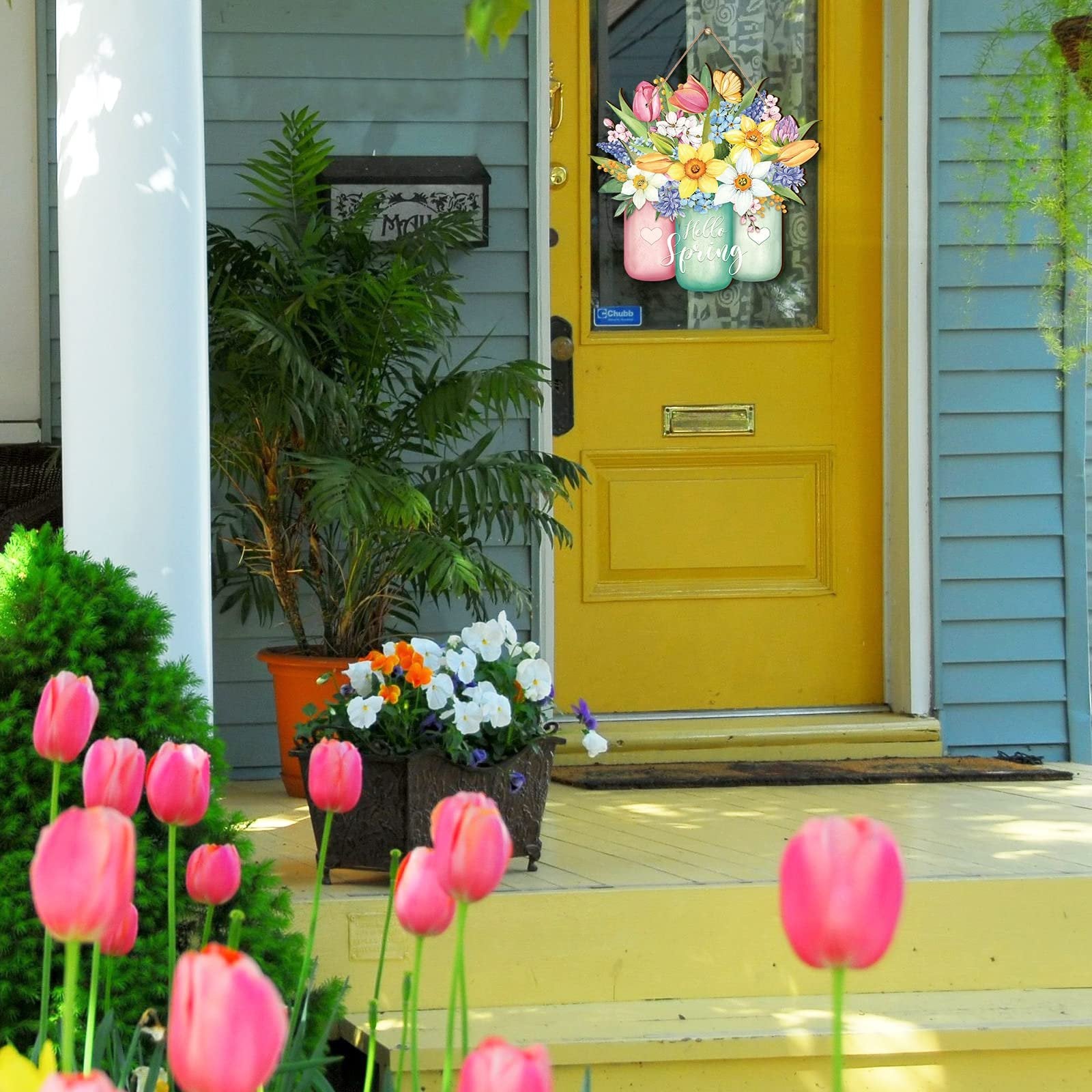 Spring door sign with flowers in jars hanging on a yellow door, with tulips and plants in the foreground.