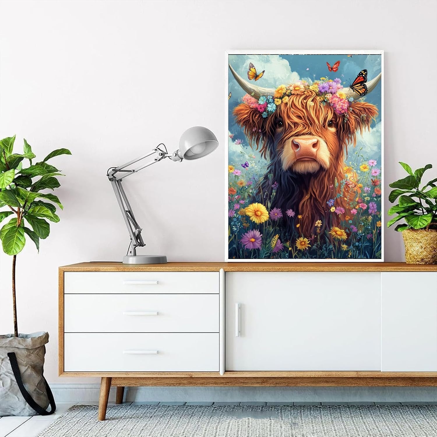 Highland cow with flowers and butterflies on canvas, displayed on a dresser with plants and a lamp.