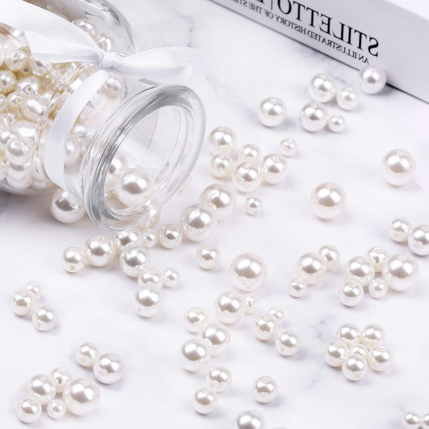Ivory pearl craft beads in assorted sizes spilling from a glass jar tied with a white ribbon.