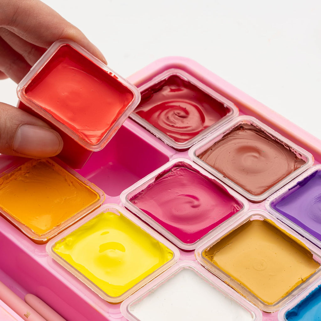 Close-up of a hand holding a red paint pot from a pink jelly gouache paint set with multiple colors visible.