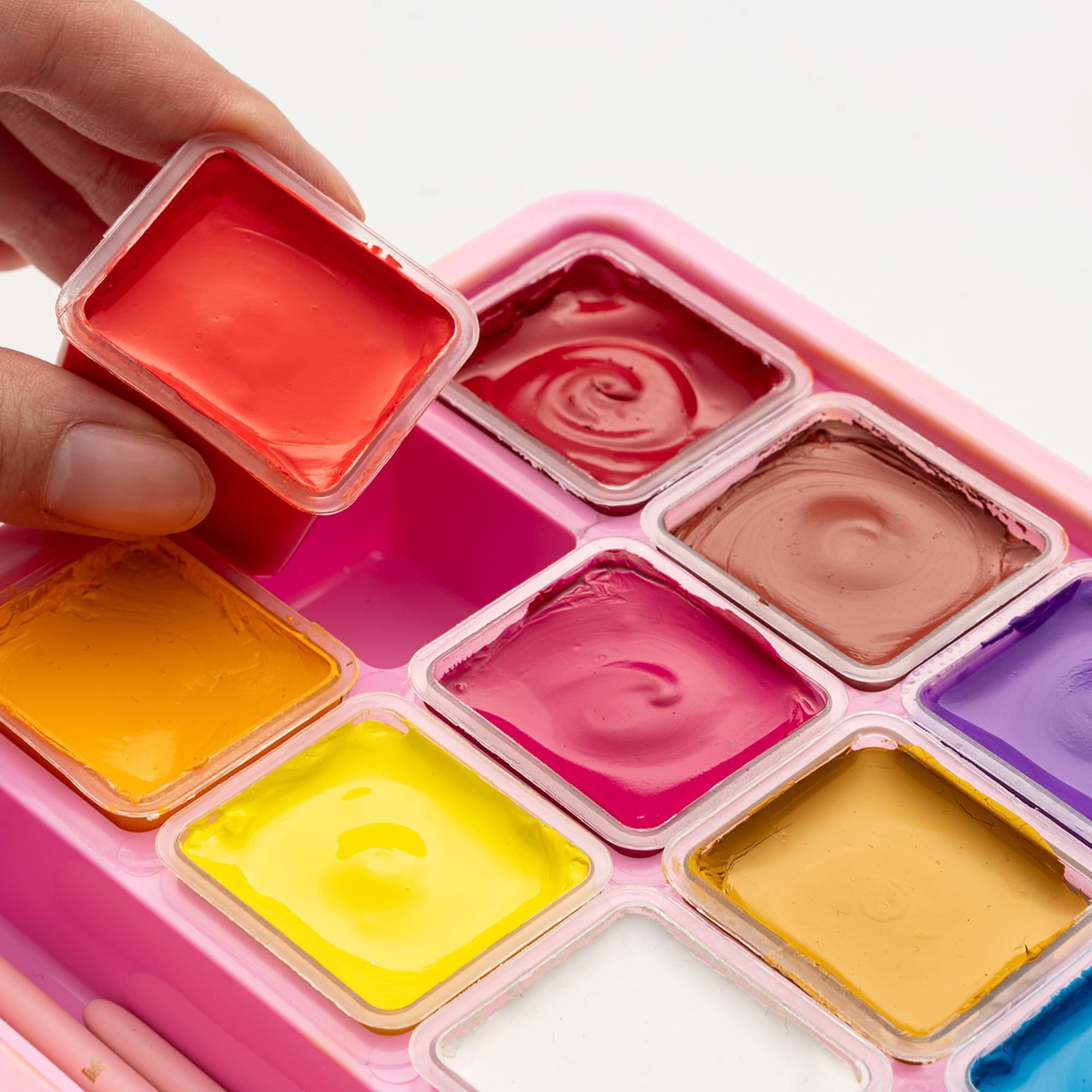 Close-up of a hand holding a red paint pot from a pink jelly gouache paint set with multiple colors visible.
