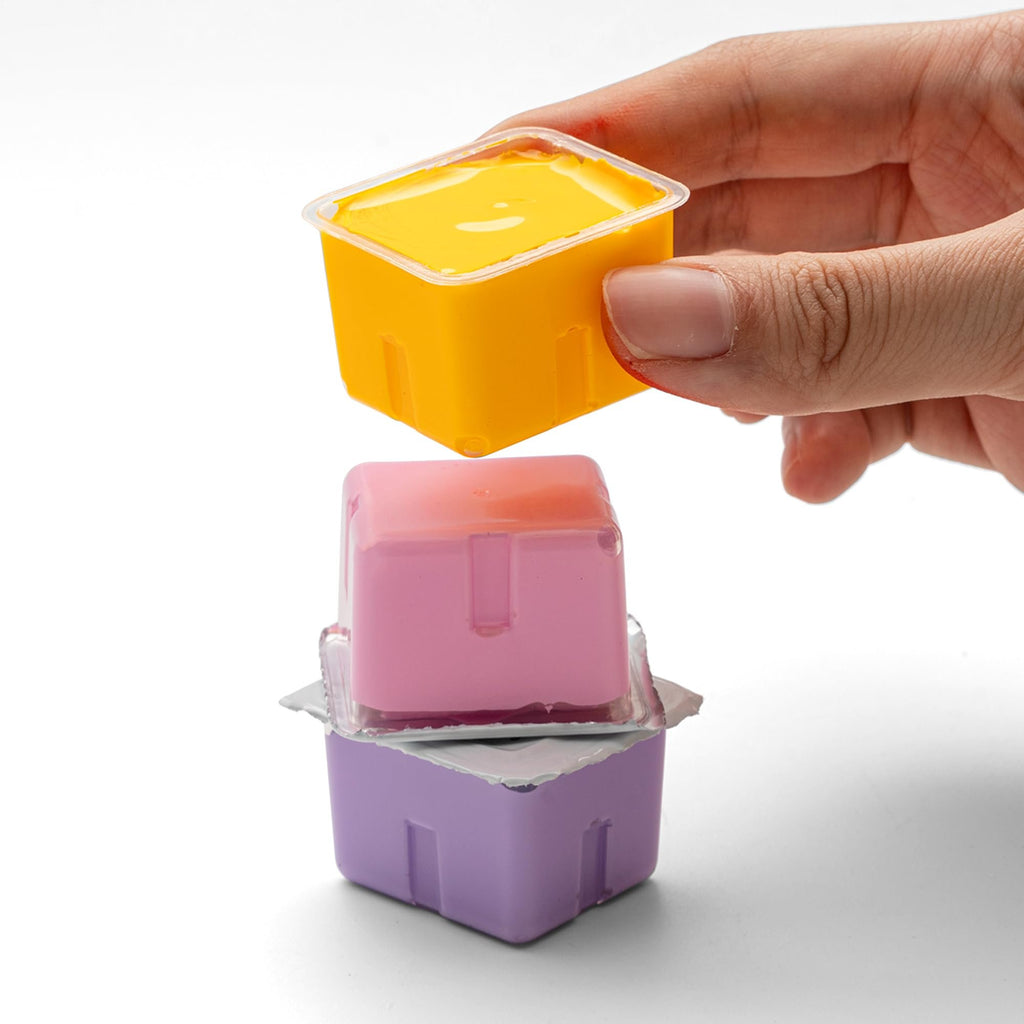 Hand holding a yellow jelly gouache paint pot above a pink one stacked on a purple one against a white background.
