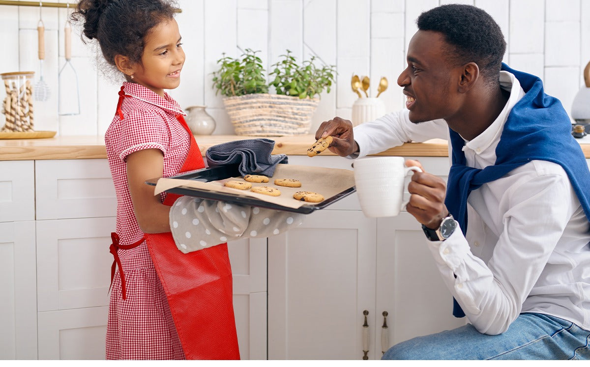 Father and daughter in kitchen, daughter wearing red apron and holding baking tray with cookies
