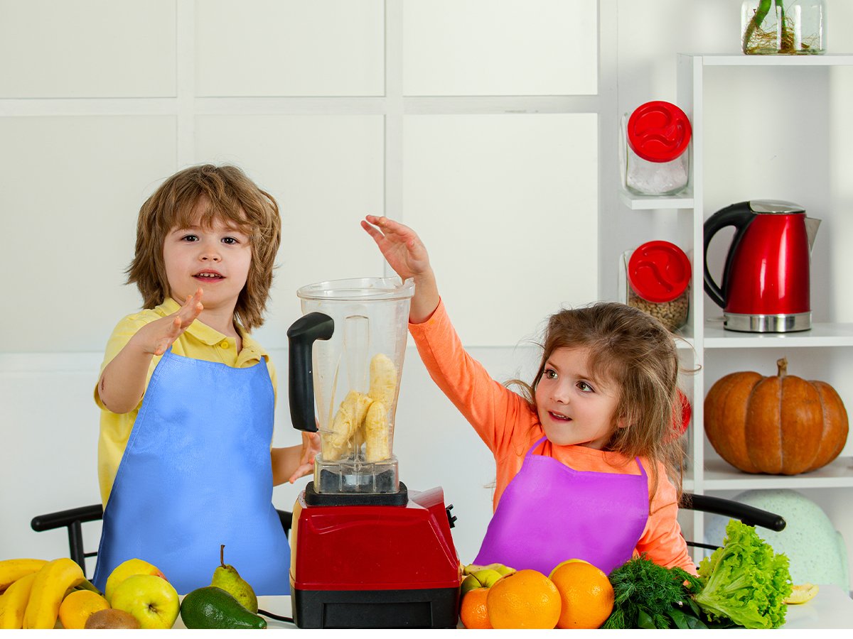 Two children wearing aprons making a smoothie in a kitchen with a blender and fruits