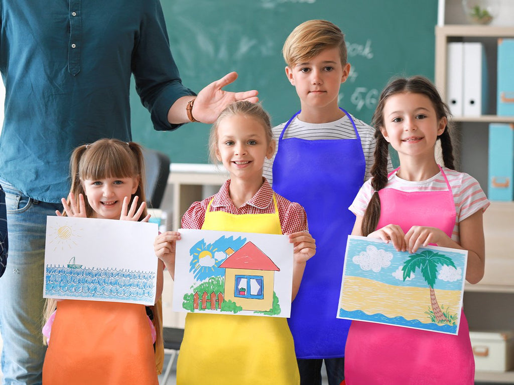 Children in colorful aprons holding up their drawings in a classroom setting with a teacher behind them.