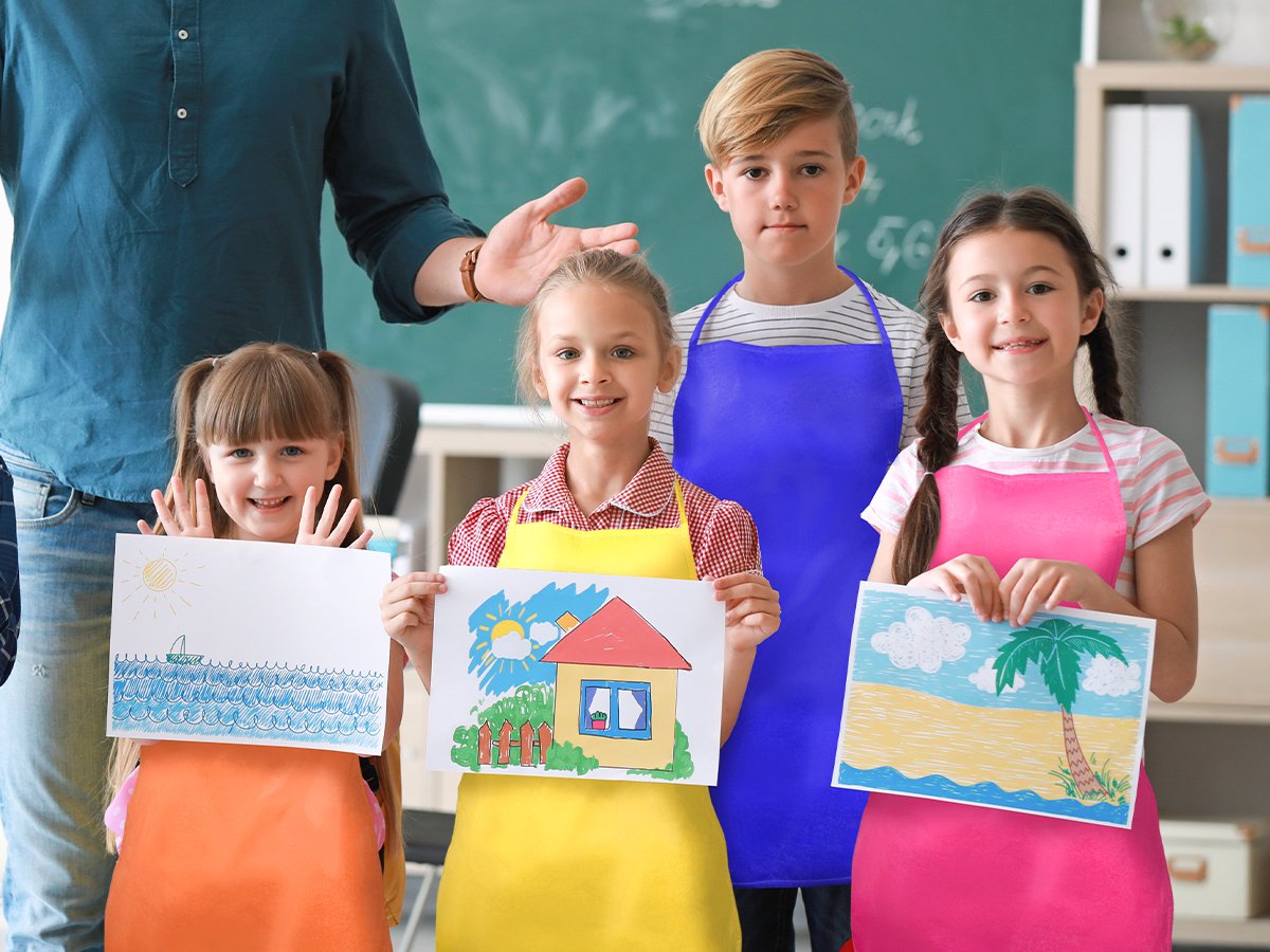 Children in colorful aprons holding up their drawings in a classroom setting with a teacher behind them.