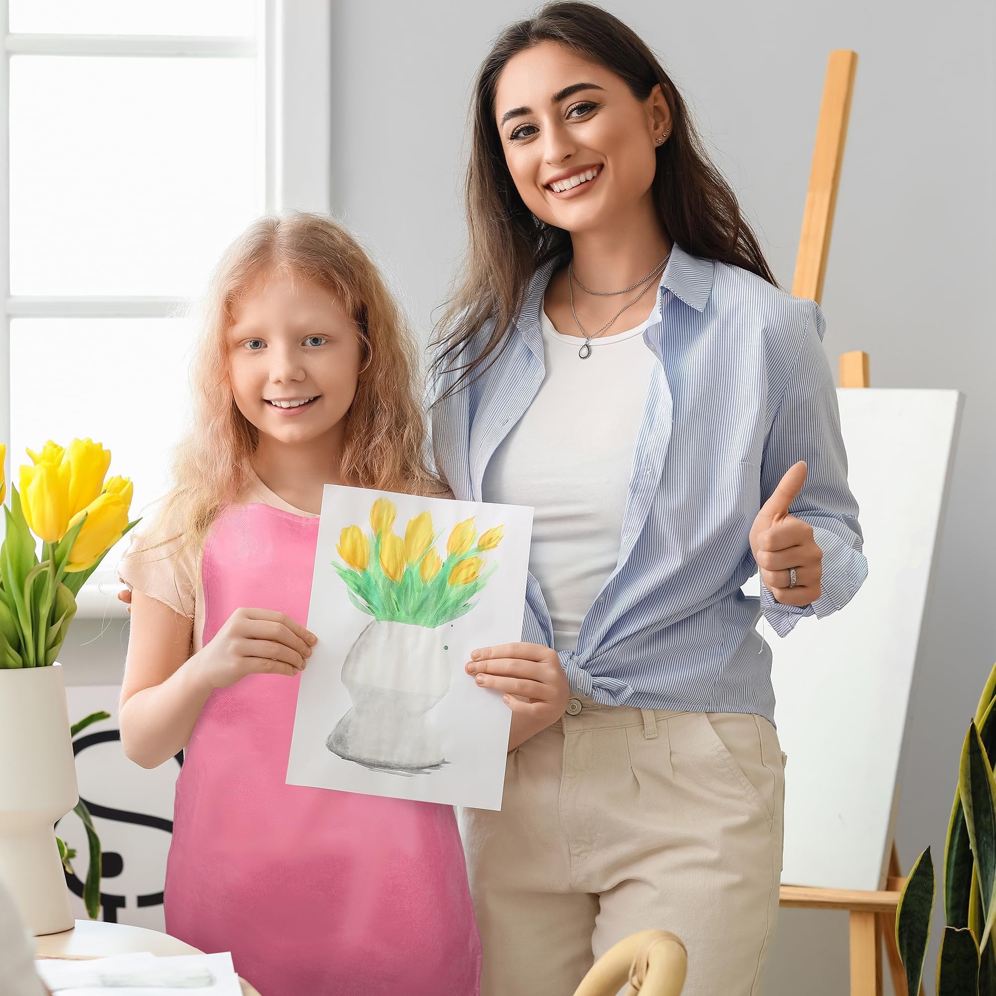 Young girl in pink apron holds tulip painting, woman gives thumbs up in art studio