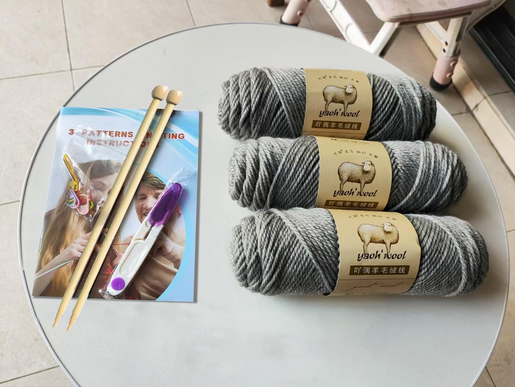 Knitting starter kit contents including three grey yarn rolls, bamboo needles, stitch markers, and instruction booklet on a white table.