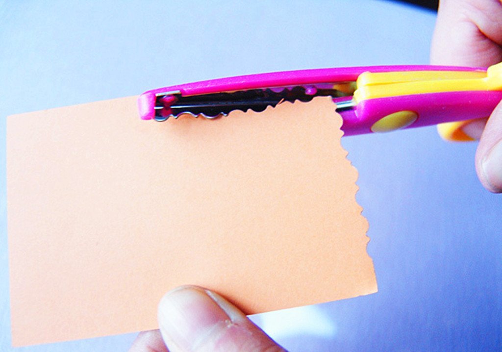 Close-up of pink and yellow craft scissors cutting a wavy edge on orange paper.