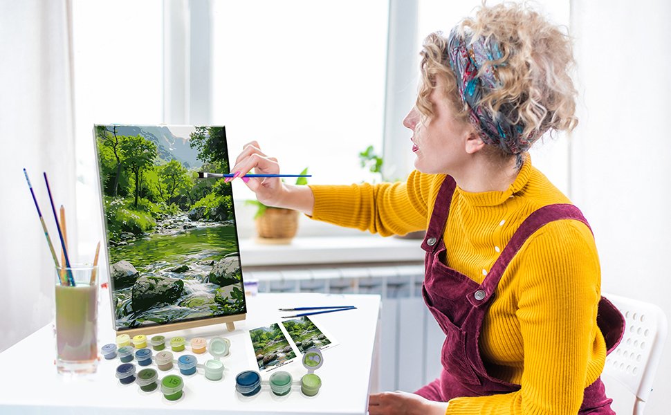 Woman painting a landscape by numbers on a canvas, with paints and brushes laid out on a table.