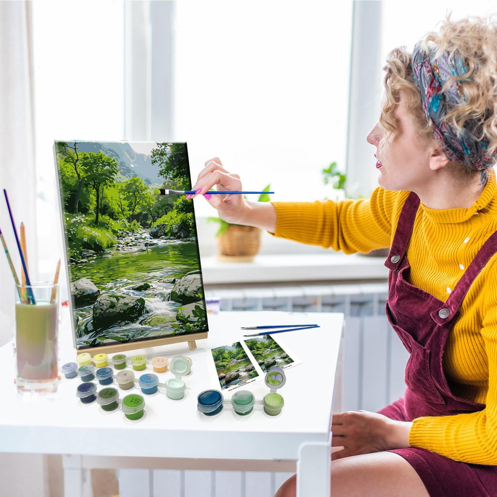 Woman painting a landscape by number kit on a canvas easel with paints and brushes on a table