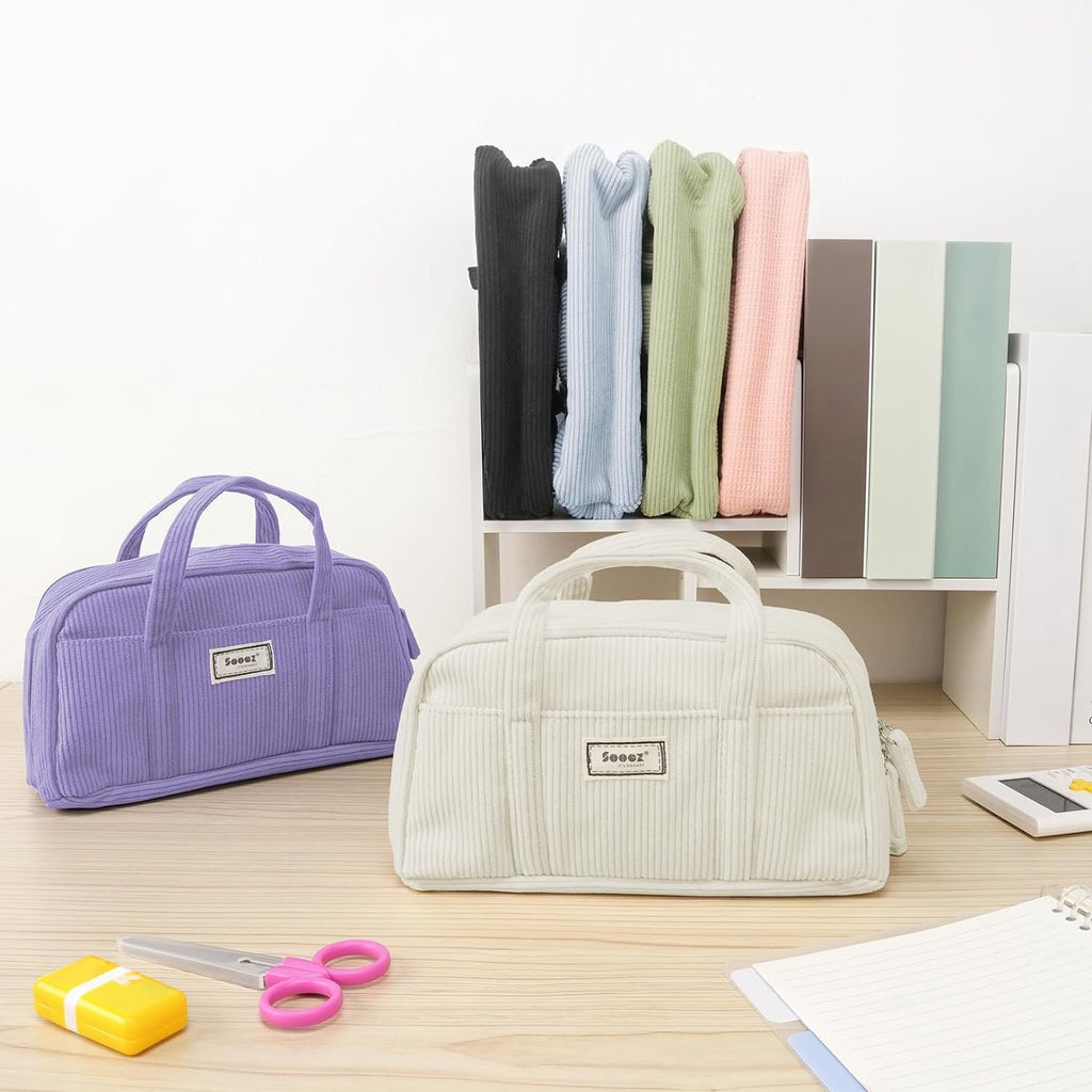 Two corduroy pencil cases, one purple and one white, on a wooden desk with stationery and colorful folded items in the background.