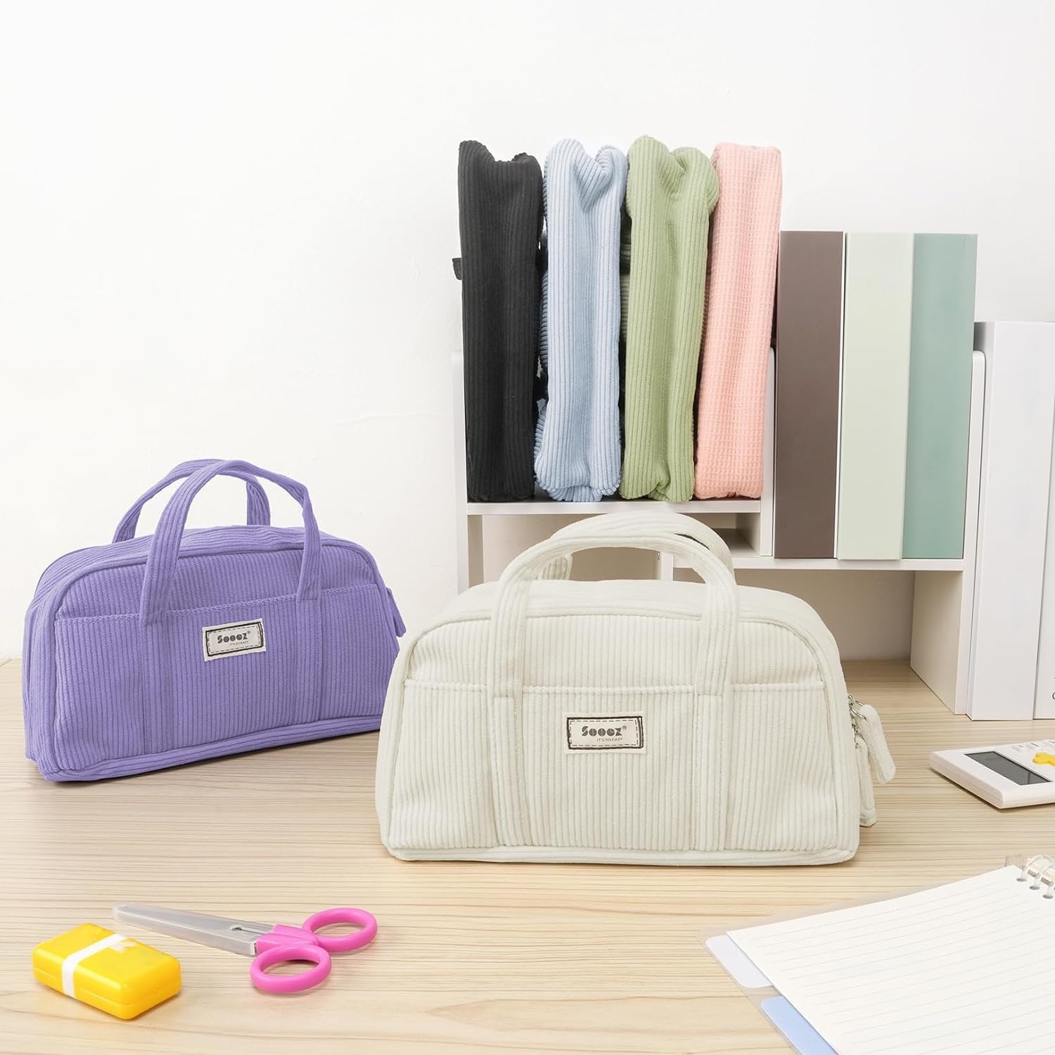 Two corduroy pencil cases, one purple and one white, on a wooden desk with stationery and colorful folded items in the background.