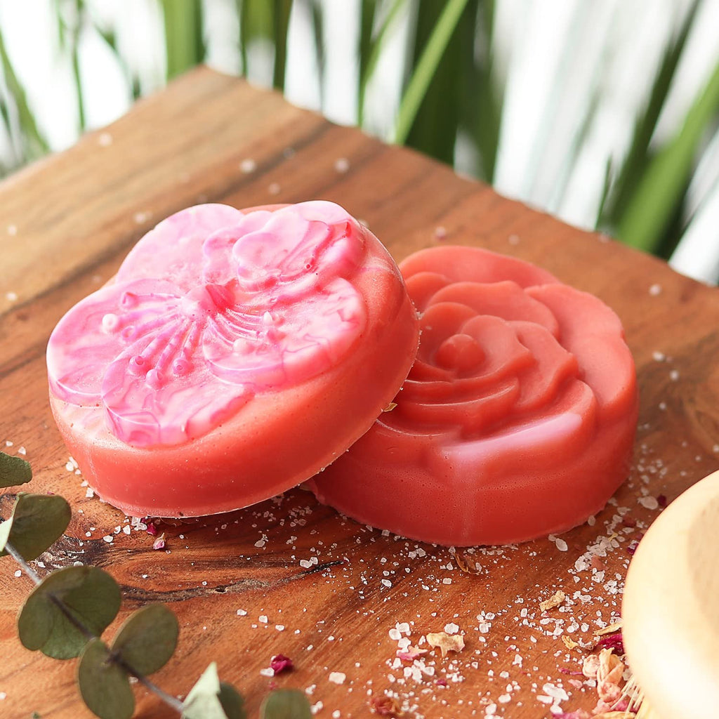 Two decorative soaps, one heart-shaped with a flower design and one rose-shaped, on a wooden board with scattered salt and dried petals.