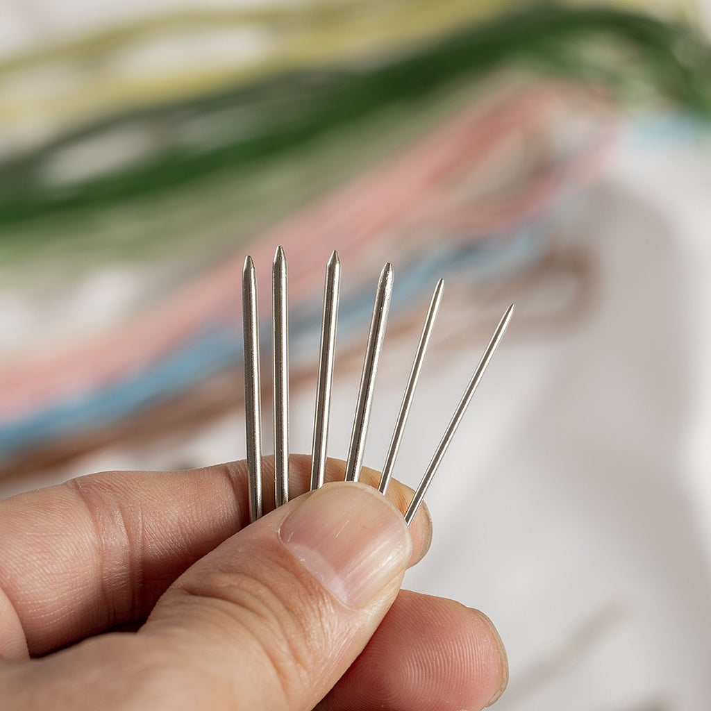 Hand holding several large eye blunt darning needles with colorful yarn blurred in the background.
