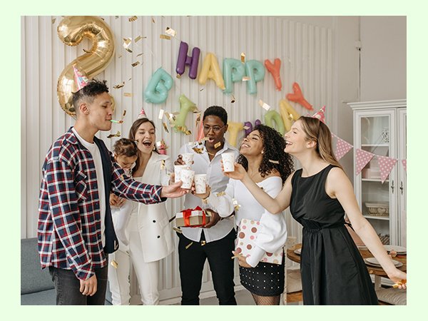Group of people celebrating a birthday with balloons and confetti, holding cups and gifts.
