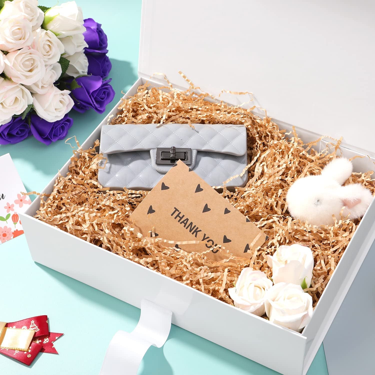Open white gift box filled with shredded paper, a gray quilted clutch, a fluffy white bunny, and white roses.