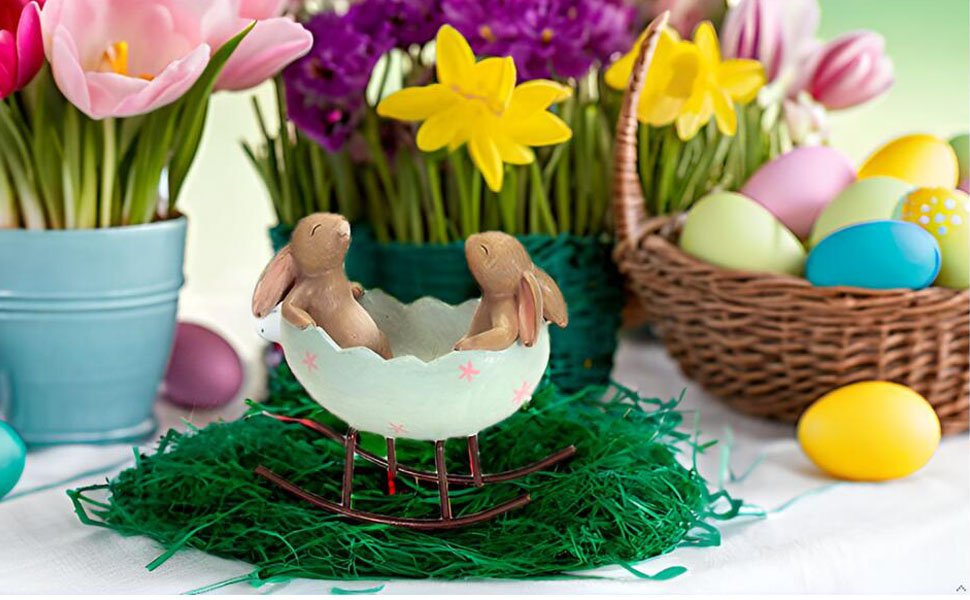Easter figurine of two bunnies in a rocking egg cradle on green grass with spring flowers and eggs